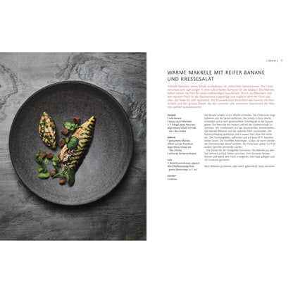 Gegrillte Makrele mit grünem Gemüse und Bananenscheiben auf dunklem Teller, daneben ein Kochbuch.