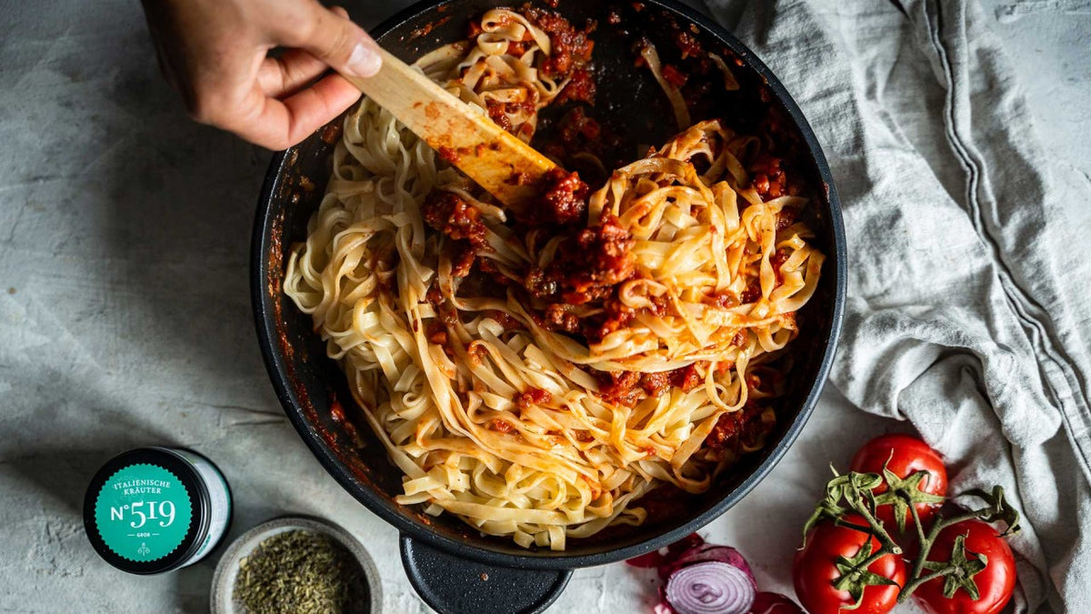 Tagliatelle mit Tomatensauce werden in einer schwarzen Pfanne mit einer Hand umgerührt, frische Tomaten liegen drum herum.