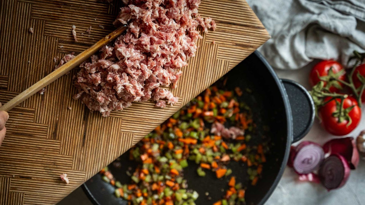 Das geschnittene Fleisch wird von einem Schneidebrett in eine Pfanne mit gewürfeltem Gemüse und Tomaten gegeben.