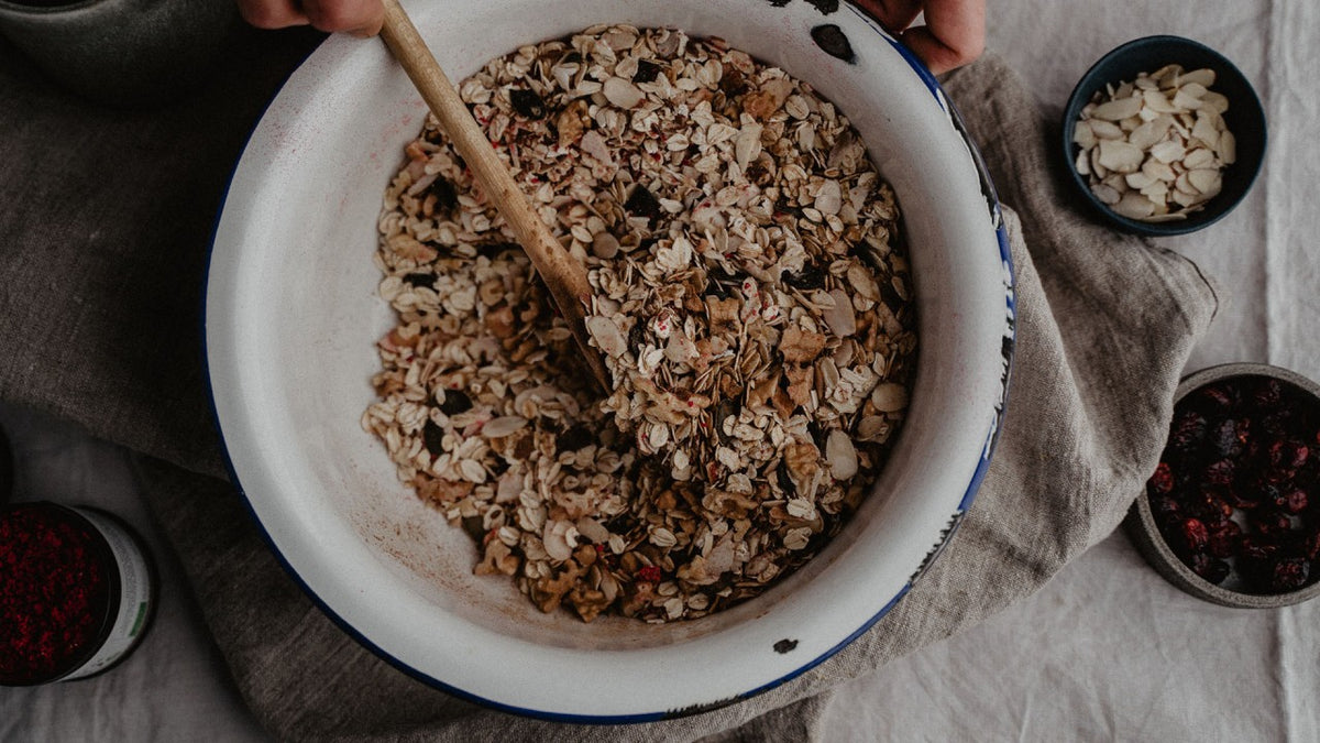 Eine Person rührt in einer Schüssel mit einem Holzlöffel Müsli auf einem mit einem Leinentuch bedeckten Tisch um.