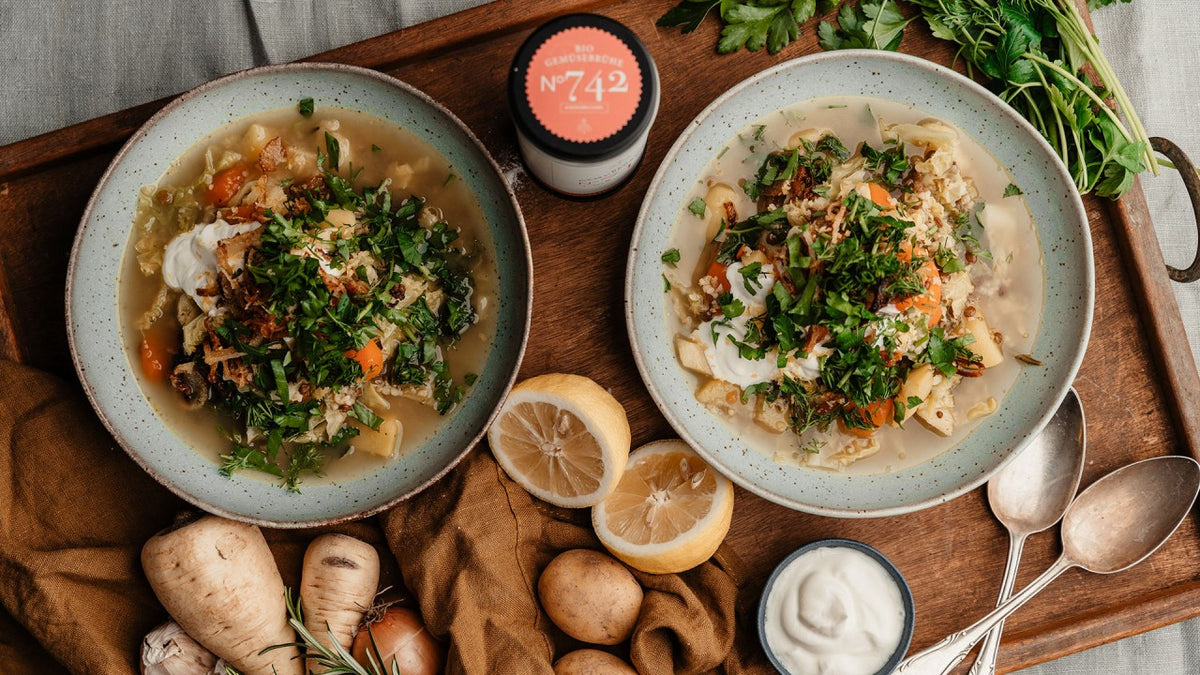 Gemüsesuppe mit Kräutern, Zitronenscheiben, Wurzelgemüse und Joghurt auf einem rustikalen Holztablett.