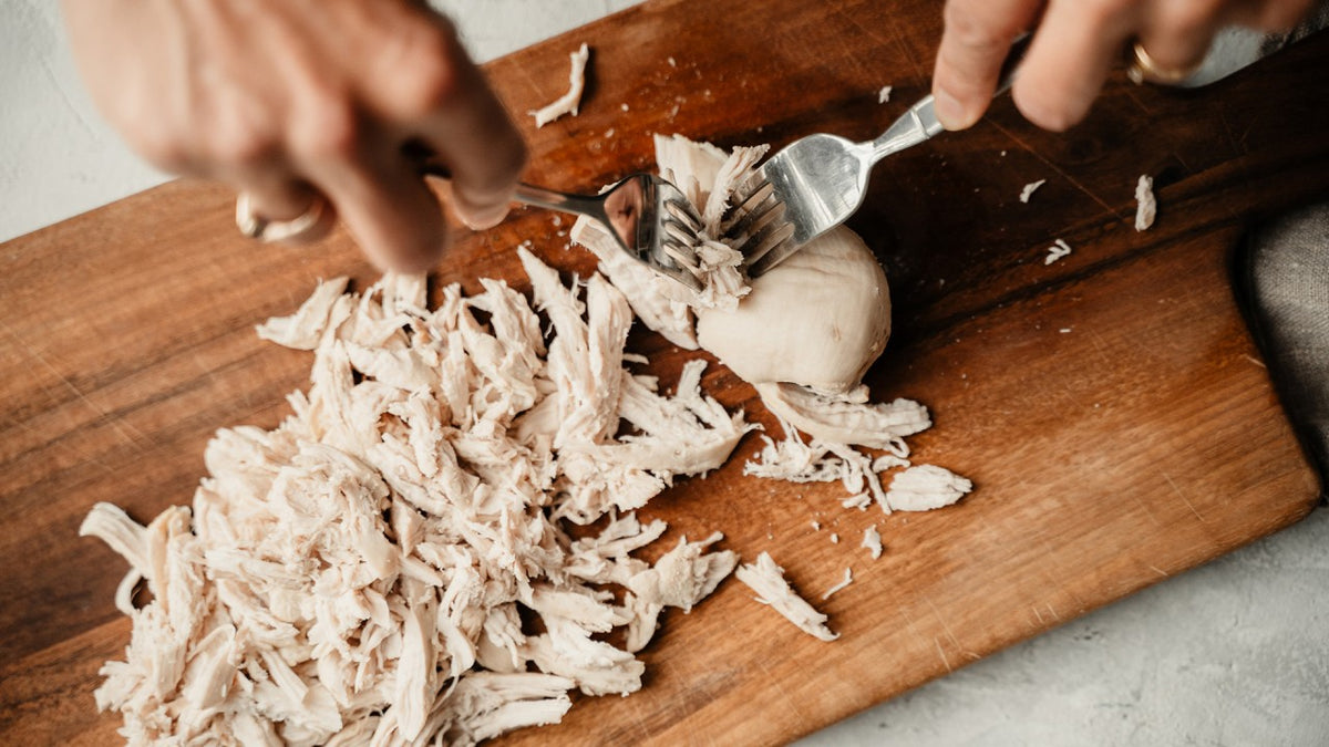 Die gekochte Hähnchenbrust wird mit Gabeln auf einem Holzbrett zerkleinert.