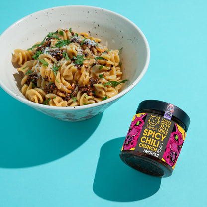 Bio Spicy Chili Crunch im Glas neben Pasta in einer Schale auf blauem Hintergrund.