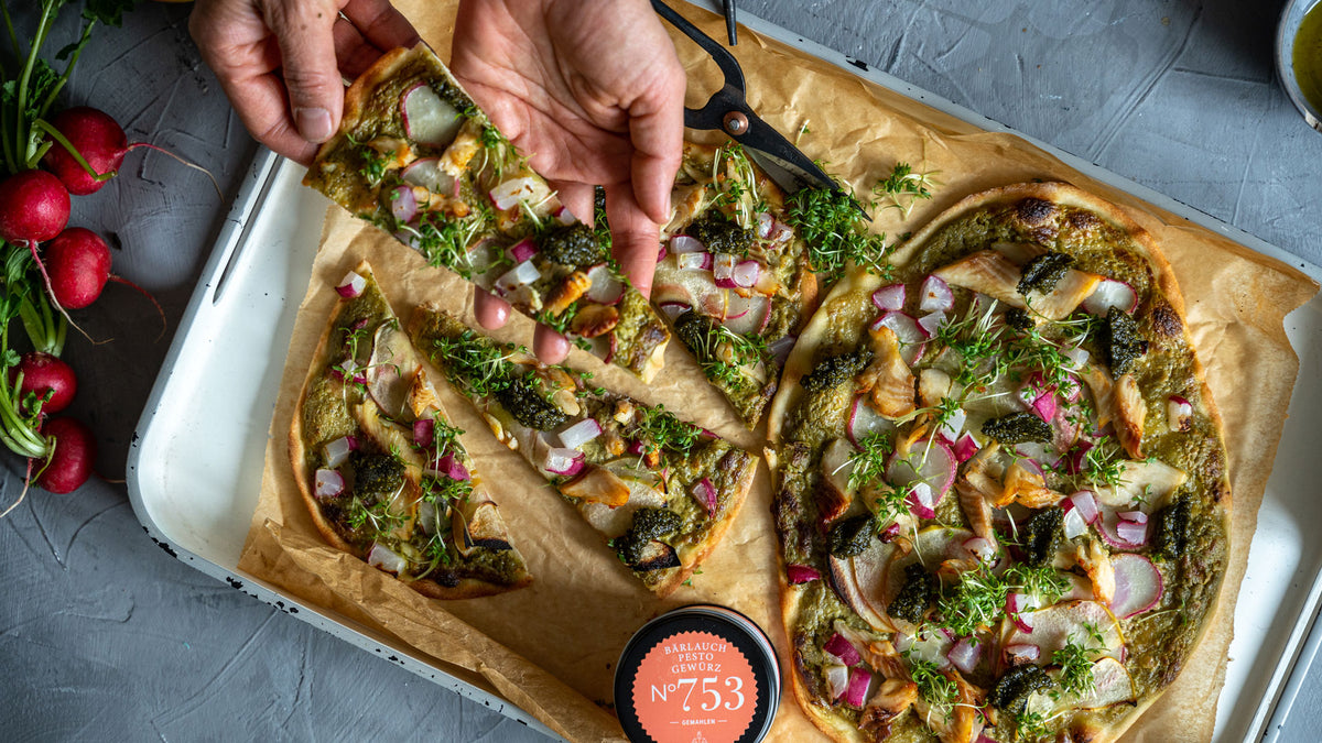 Hände halten ein Stück Gemüsepizza, daneben frische Radieschen und Pesto-Gewürze auf Backpapier.
