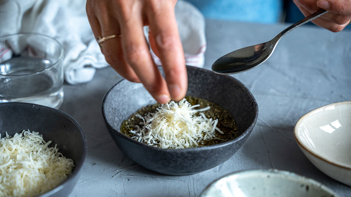 Eine Hand streut geriebenen Käse auf eine Schüssel mit Pesto-Gewürzen, daneben liegt ein Löffel.
