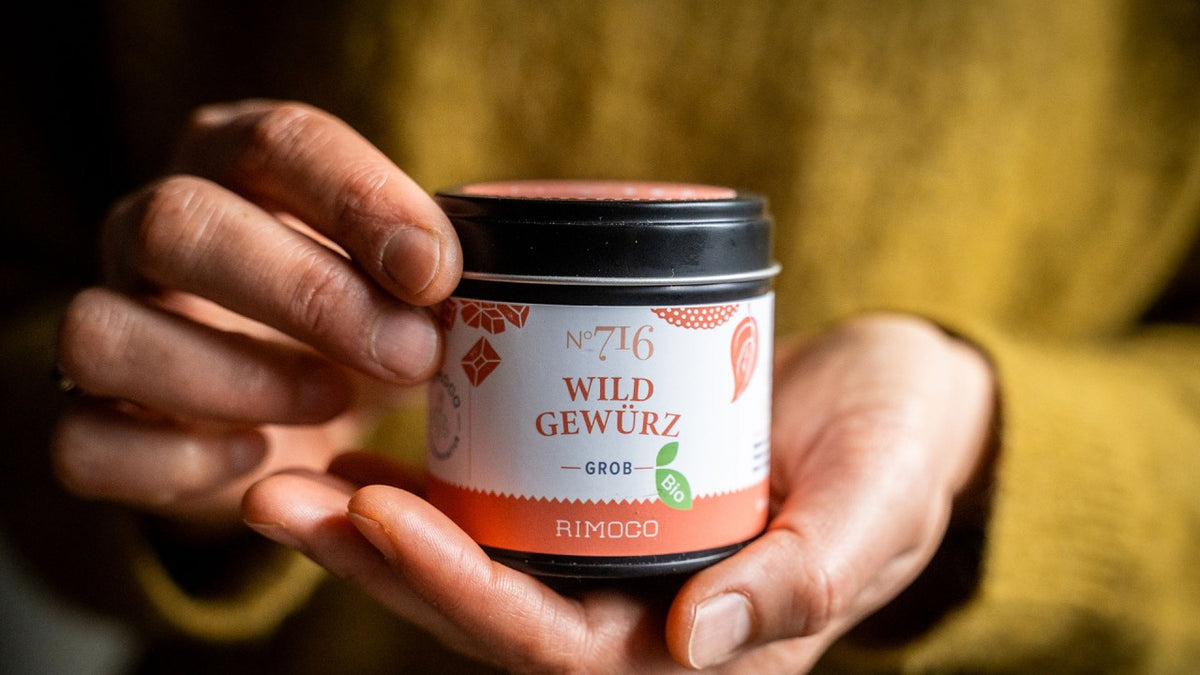 Eine Person in einem gelben Pullover hält mit beiden Händen eine kleine Dose mit der Aufschrift „Wild Gewürz“ in der Hand.