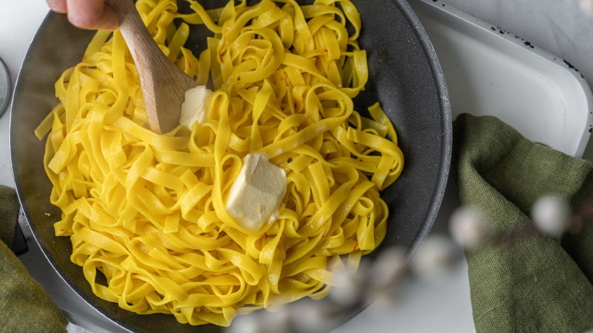Gelbe Tagliatelle und Butter werden mit einem Holzlöffel in einer schwarzen Pfanne verrührt.