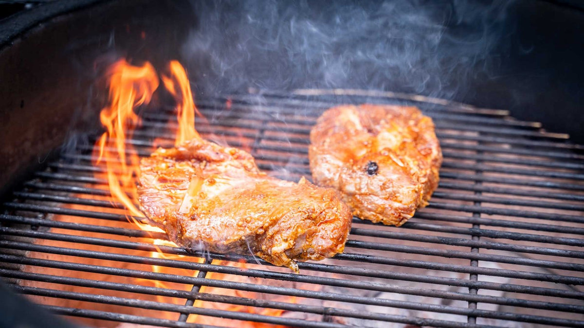 Steaks brutzeln auf dem glühenden Grill, Rauch und Flammen steigen um die Fleischstücke herum auf.