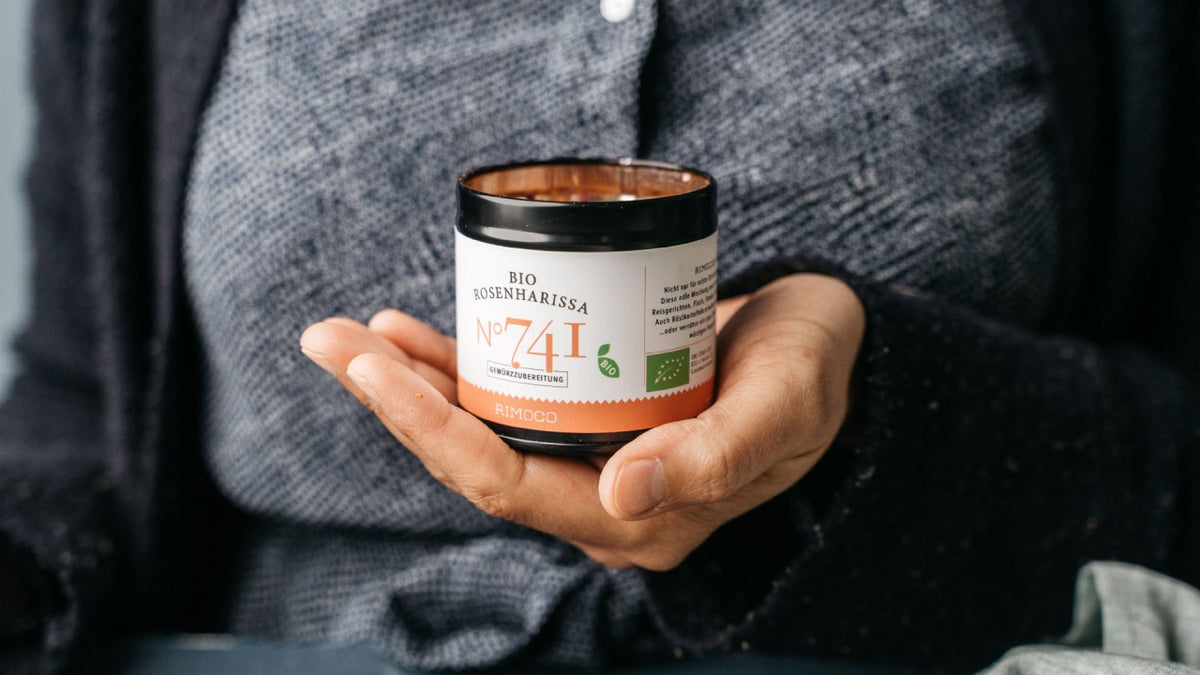 Eine Person hält ein Glas Bio-Rosenharissa Nr. 741 mit deutschen und englischen Beschriftungen in der Hand, eine Gewürzmischung.