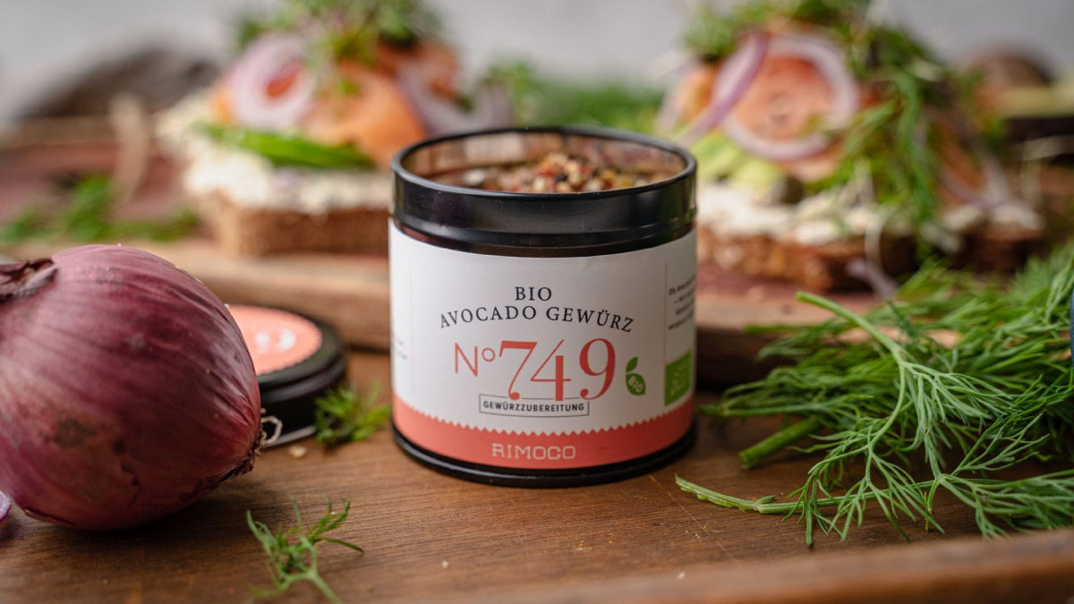 Ein Avocado-Gewürzglas auf einem Holzbrett, daneben sind Dill und belegte Brotscheiben zu sehen.