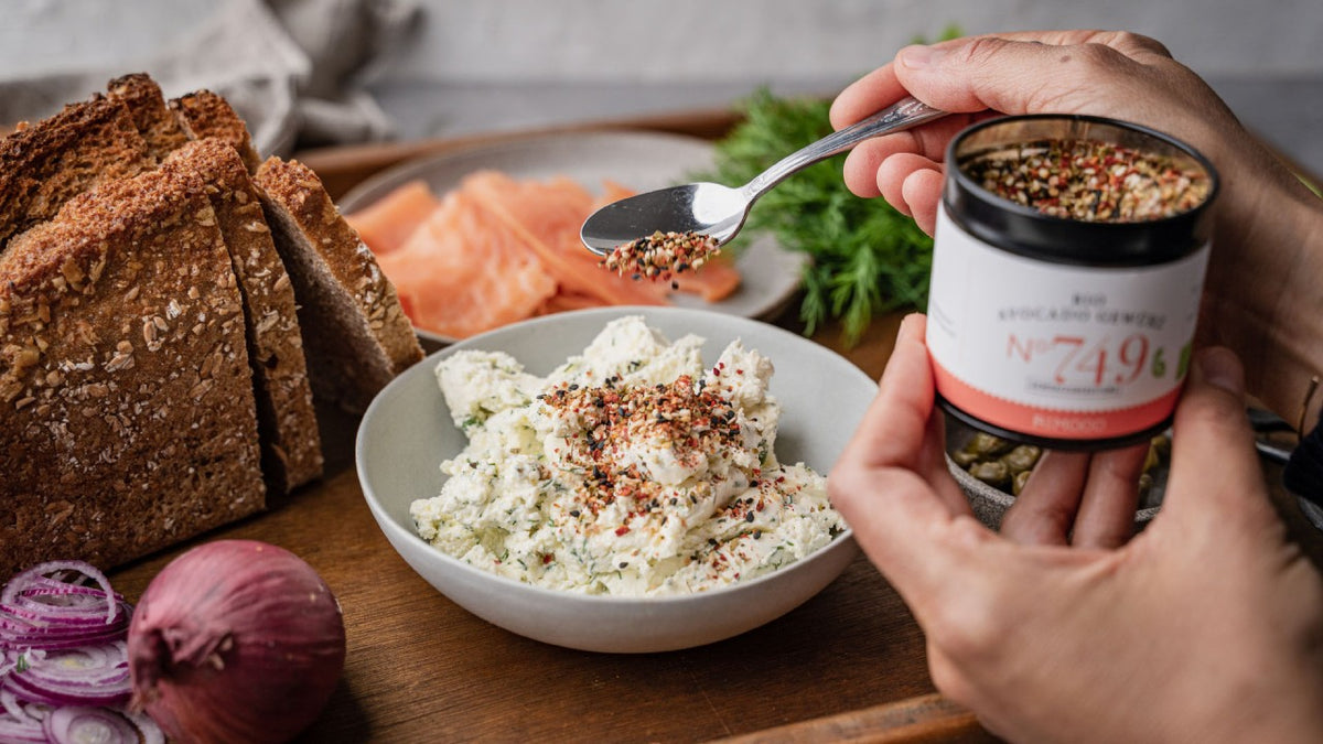 Brot, Räucherlachs und Zwiebeln mit Frischkäse auf einem Holztablett, darüber werden Gewürze gestreut.