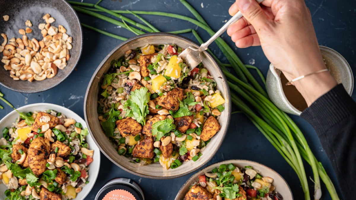 Eine Hand hält eine Gabel über einem bunten Salat mit Tofu, Mango, Erdnüssen, Koriander und Gemüse.