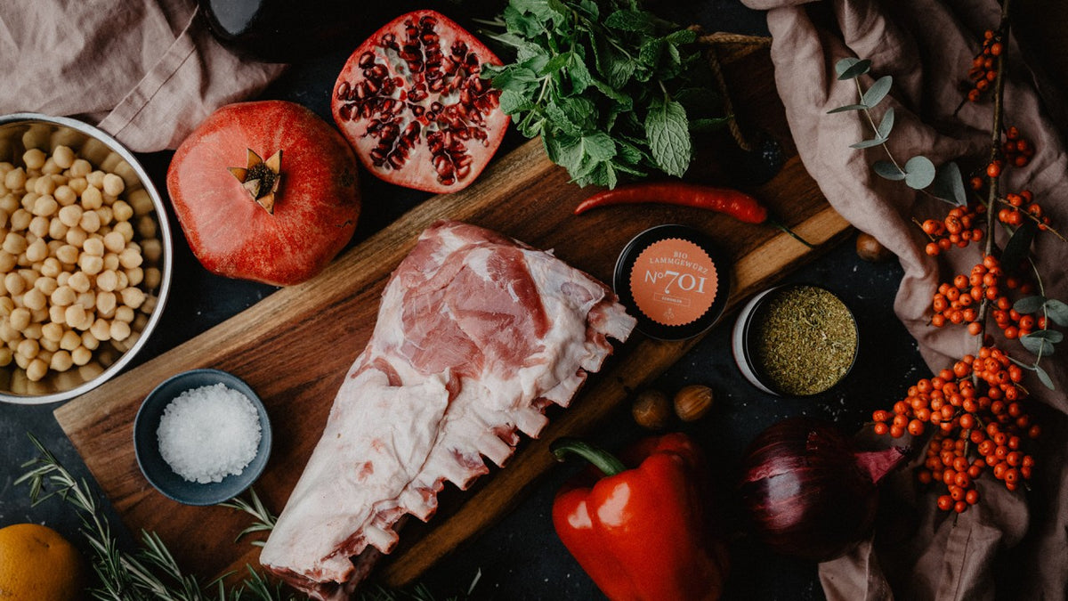 Rohes Lammfleisch, Granatapfelkerne, Kichererbsen und frische Kräuter auf einem Holzbrett.