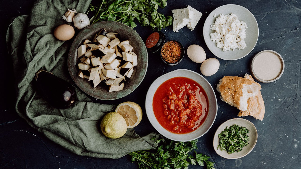 Aubergine, Eier, Feta, Kräuter, Zitrone, Tomatensauce und Knoblauch auf dunklem Hintergrund.