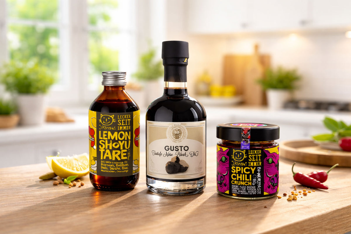 Flasche Lemon Shoyu Tare, schwarze Balsamico Sauce und Glas Spicy Chili Crunch auf Holztisch mit Zitrone und Chili in heller Küche