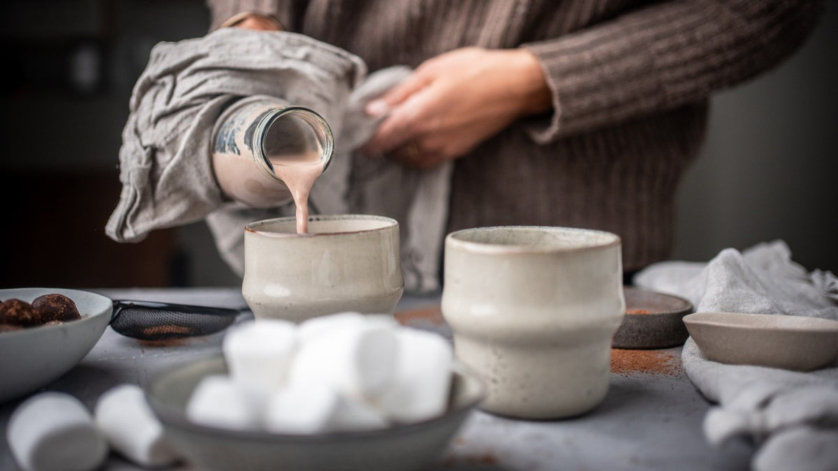 Jemand gießt Kakao in eine Keramiktasse, daneben liegen Marshmallows und eine weitere Tasse.