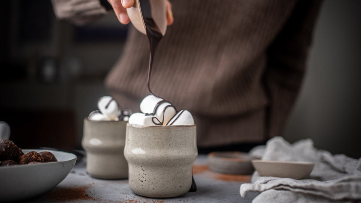 Schokoladensoße über Marshmallows in Tassen mit Kakao auf einem gemütlichen Tisch.