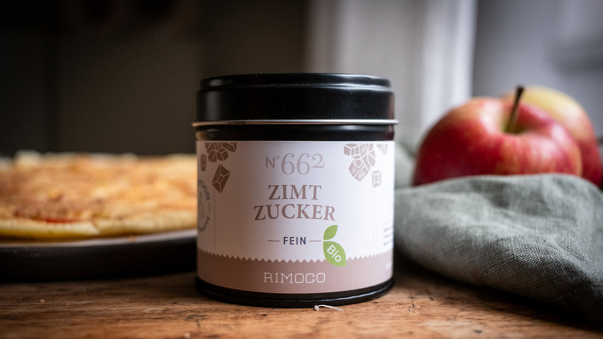 Ein mit Zimt bestäubtes Gebäck liegt neben einem Apfel und einer Dose mit der Aufschrift „Zimt-Zucker“.