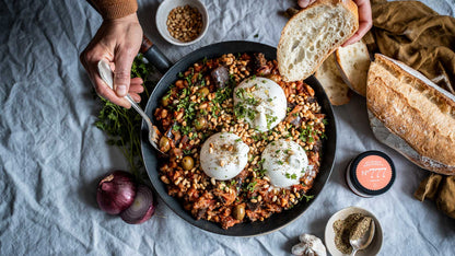 Shakshuka mit Eiern, Kräutern und Pinienkernen; eine Hand tunkt Brot hinein, rustikaler Tisch.