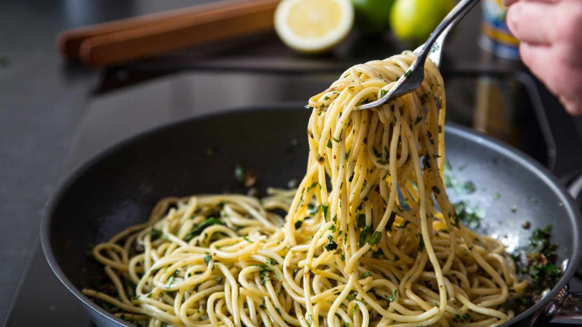 Eine Gabel hebt Spaghetti mit Kräutern aus einer Pfanne, im Hintergrund sind eine Zitrone und Besteck unscharf zu sehen.