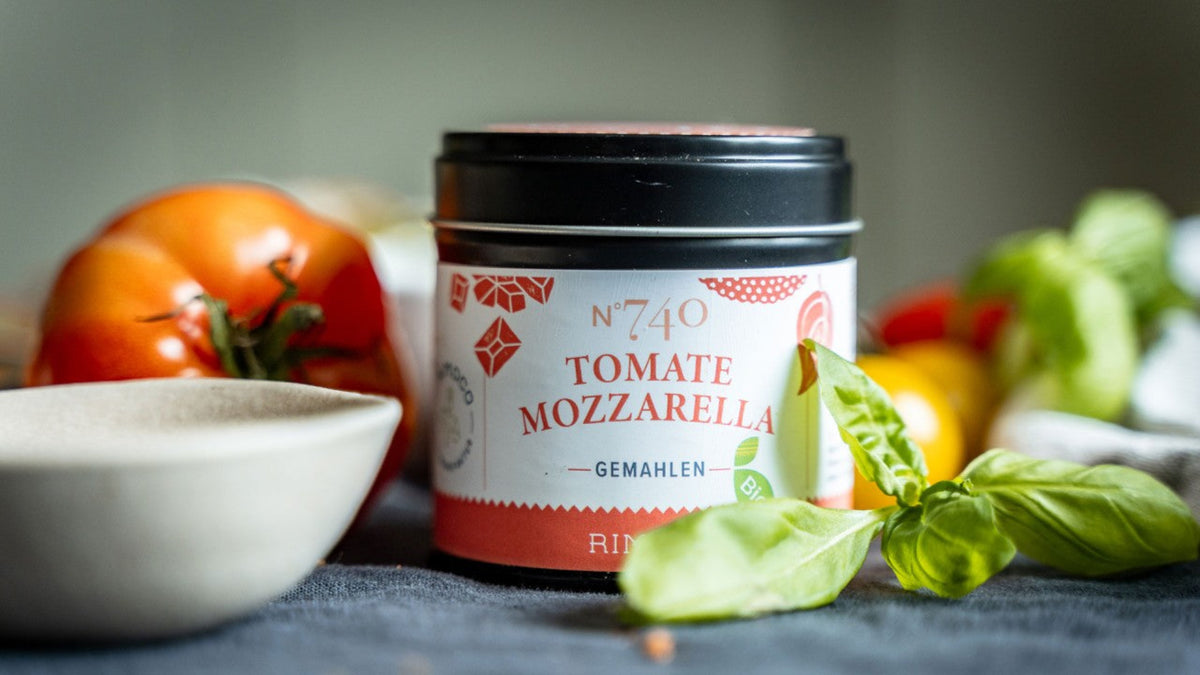 Ein Gewürzglas mit der Aufschrift „Tomate Mozzarella“ steht neben frischen Tomaten und Basilikumblättern.