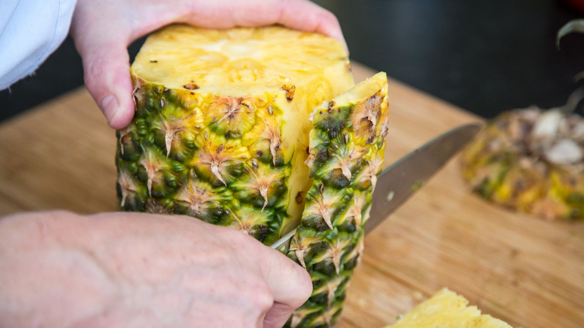 Mit einem Messer schneiden die Hände auf einem Holzbrett die Schale einer reifen Ananas auf.