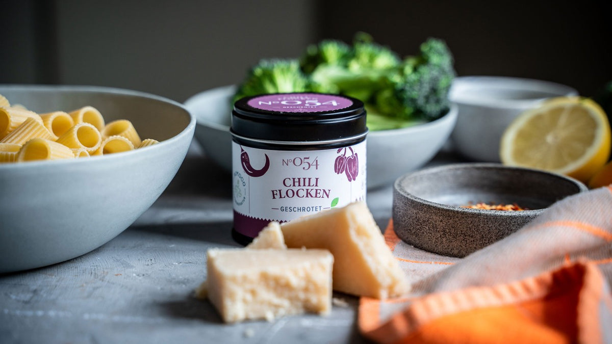 Ein Glas Chiliflocken neben Nudeln, Käse, Brokkoli, Zitrone und weiteren Gewürzen.