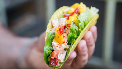Eine Hand hält einen Taco mit Fisch, Salat, Mango, Tomaten, Pico de Gallo und gemahlenem grünen Chili.