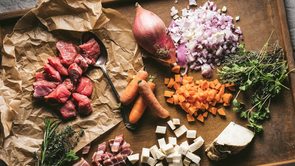Geschnittenes Gemüse, Kräuter und rohes Fleisch mit Gewürzen auf einem Holzbrett und Backpapier.
