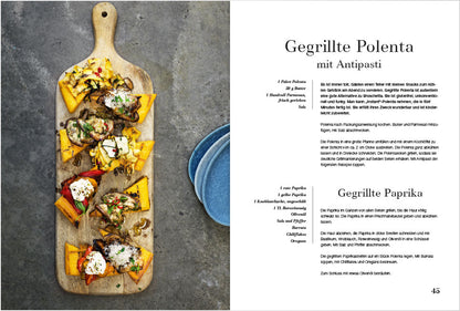 Gegrillte Polenta und Antipasti auf Holzbrett, daneben das Grillgemüse Kochbuch auf Deutsch.