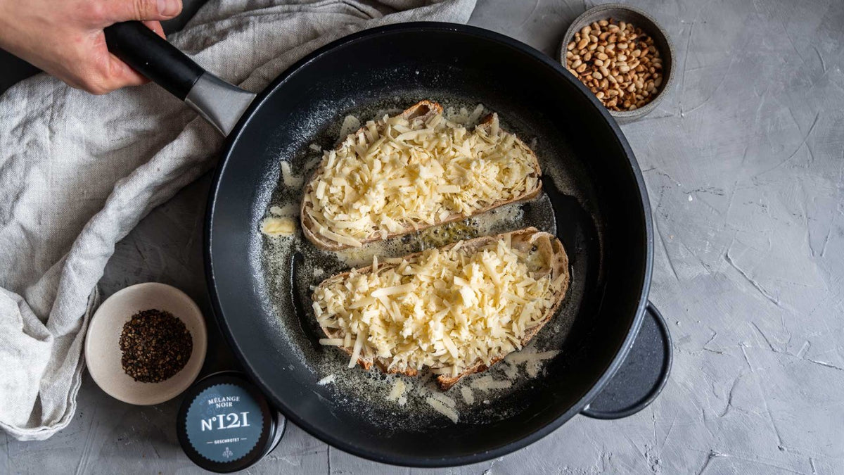 Zwei Brotscheiben mit geriebenem Käse, groben Gewürzen und Pinienkernen in einer Pfanne.