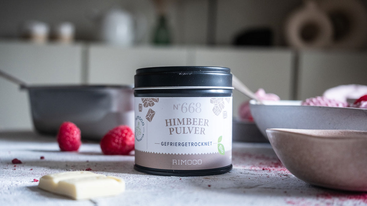 Dose mit Himbeerpulver auf dem Tisch, daneben frische Himbeeren, weiße Schokolade und Schalen.