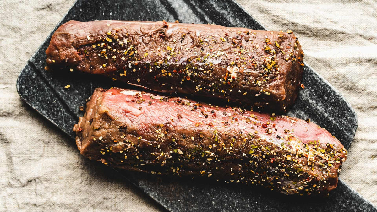 Zwei rohe, gewürzte Steaks auf einem dunklen Brett auf hellem Stoff.