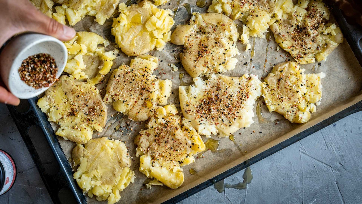 Zerkleinerte Kartoffeln auf Backpapier, eine Hand streut gemahlene Gewürze darüber.