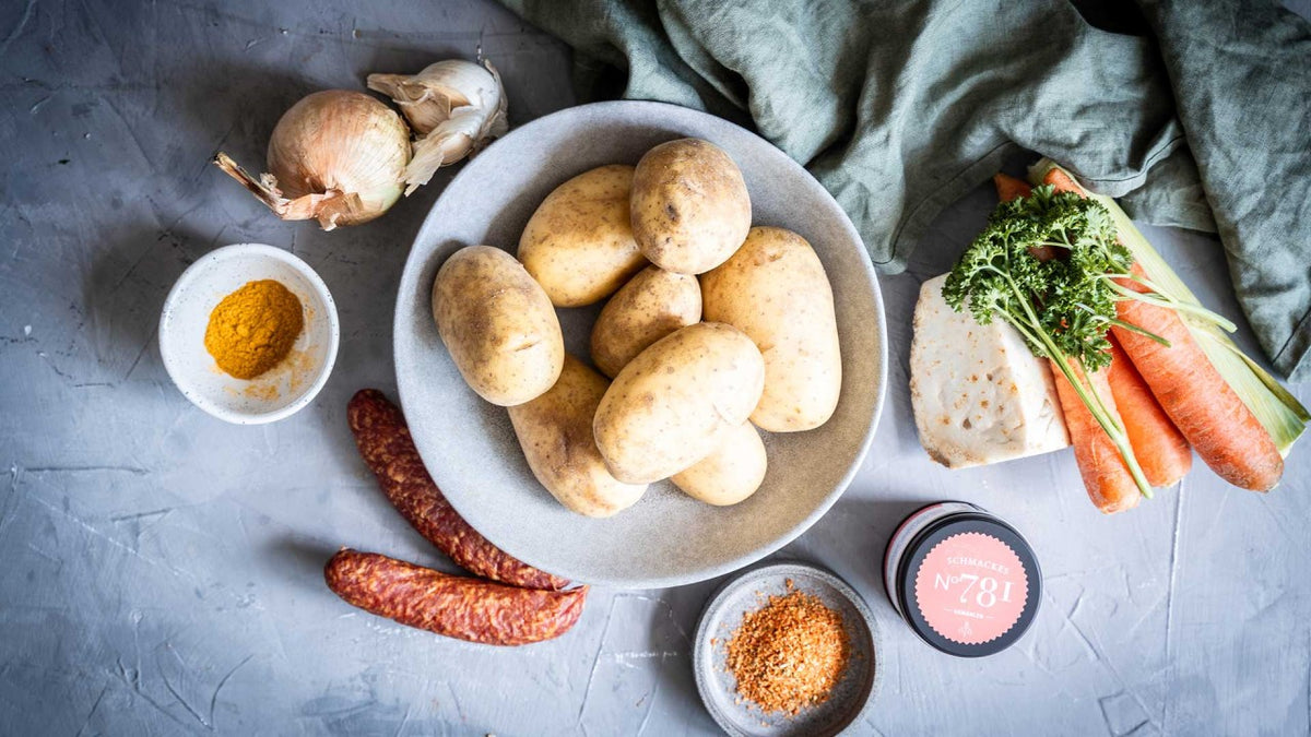 Eine Schüssel mit Kartoffeln, Karotten, Zwiebeln, Knoblauch, Wurst, Gewürzen und frischen Kräutern auf grauem Hintergrund.