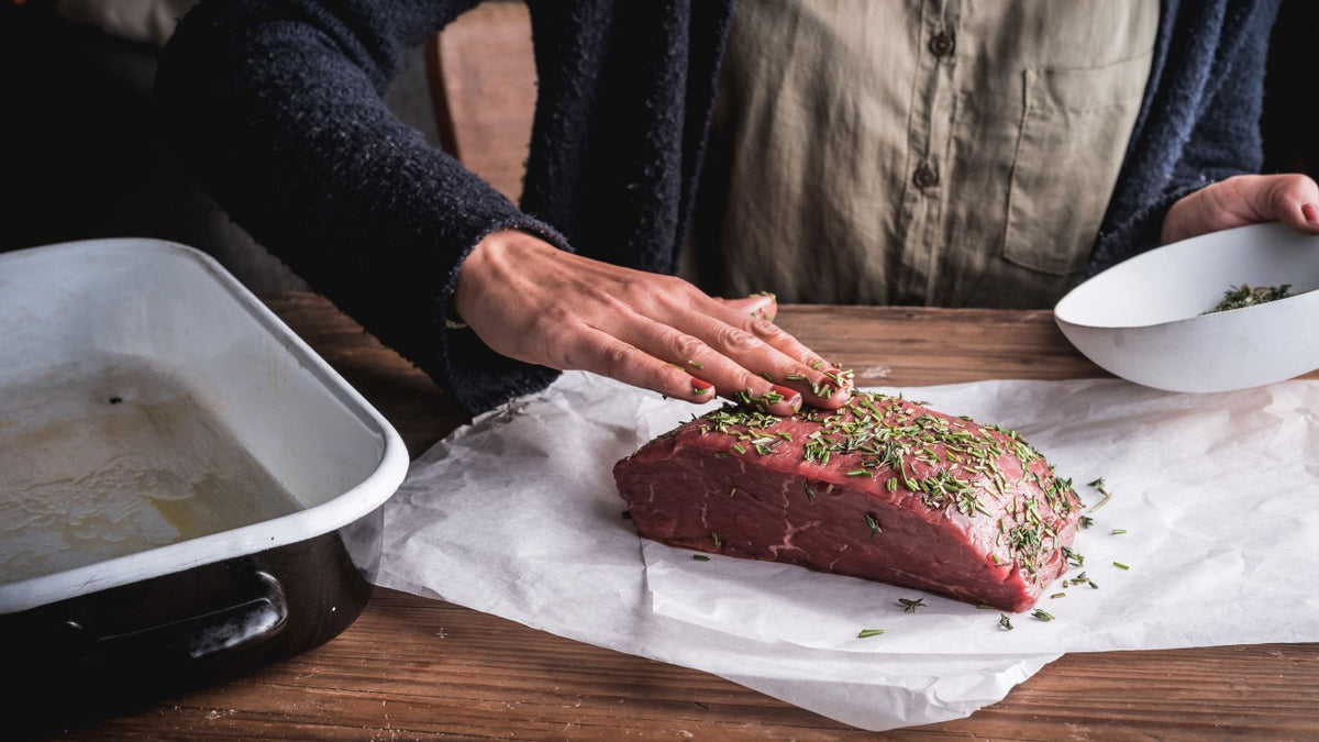 Eine Person würzt einen rohen Rinderbraten mit frischen Kräutern auf Backpapier neben einer Bratpfanne.