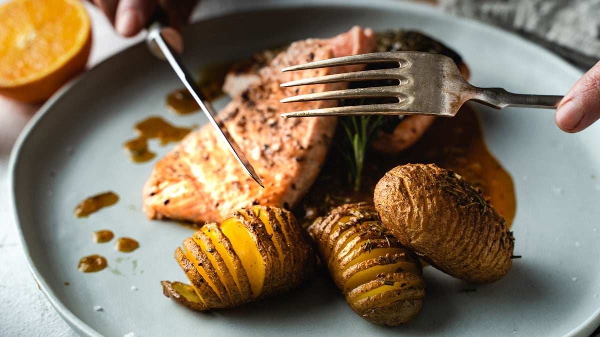 Ein Teller mit gebackenem Lachs, Hasselback-Kartoffeln, cremiger Soße und einer Gabel.