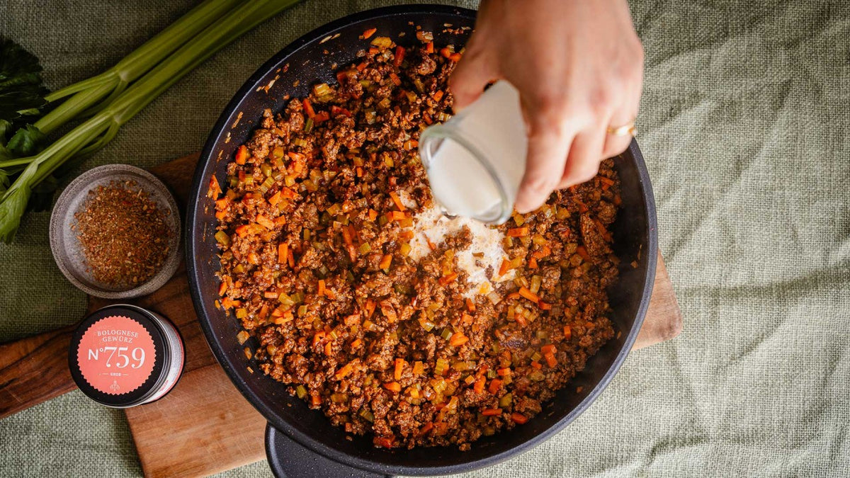Milch wird in eine Pfanne mit Hackfleisch, Karotten und sichtbaren groben Gewürzen auf einem Holzbrett gegossen.