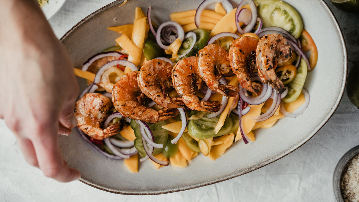 Cajun-Garnelen auf Mangosalat mit roten Zwiebeln, eine Hand greift zum Teller.