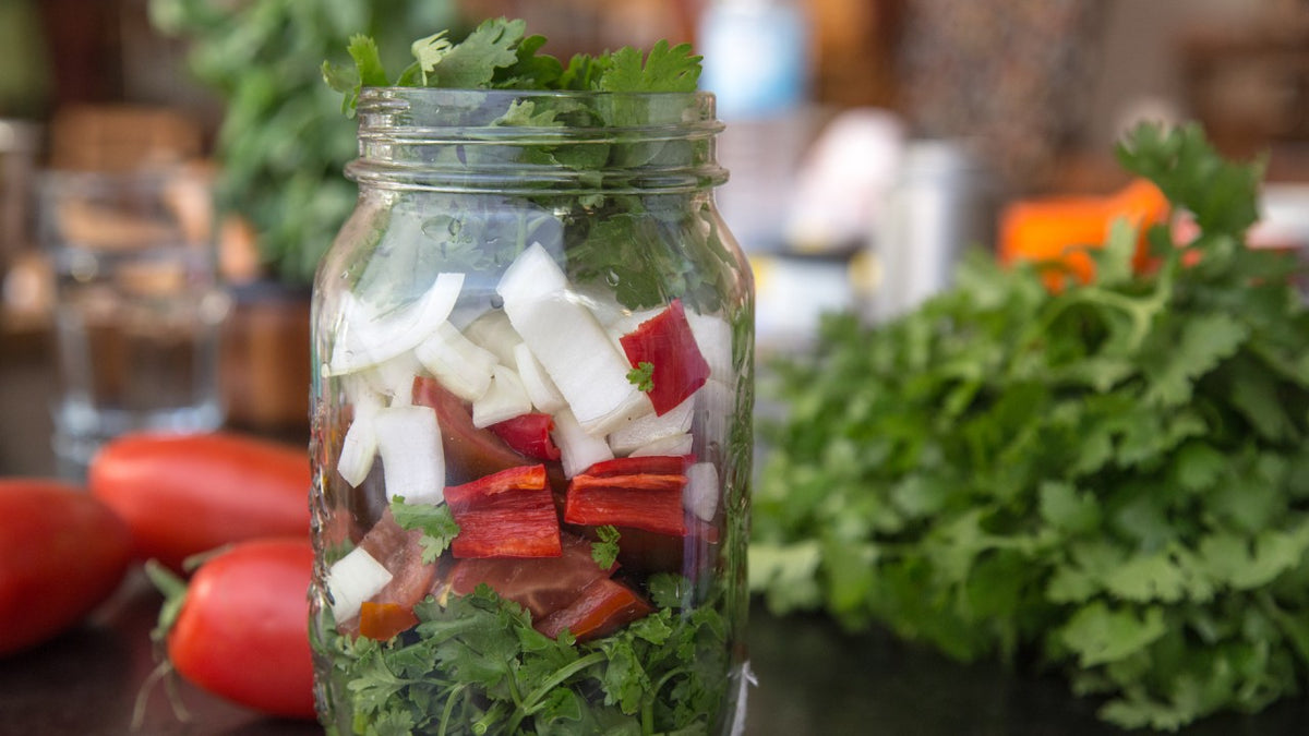Ein Einmachglas mit gehackten Zwiebeln, roten Paprikaschoten, Tomaten und frischem Koriander auf einer Theke.