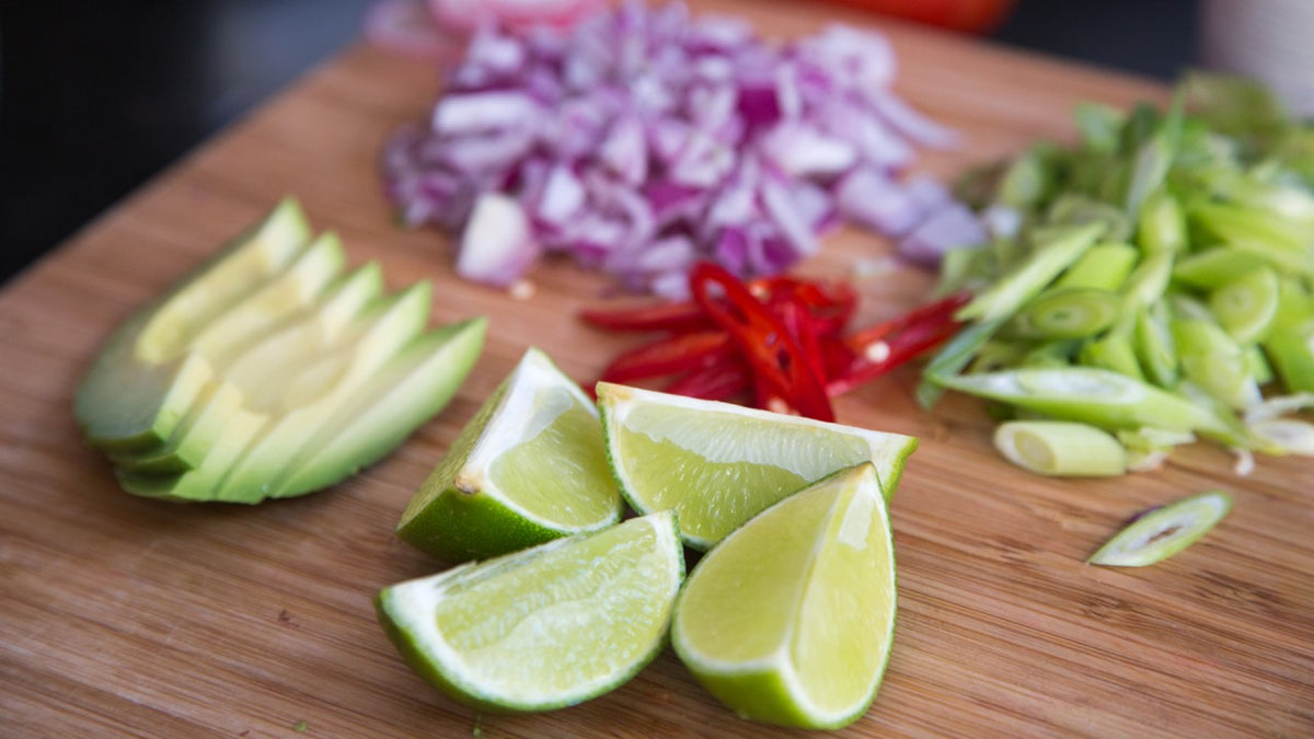 Geschnittene Limette, Avocado, rote Chilischote und Frühlingszwiebeln, auf einem Holzbrett angerichtet.