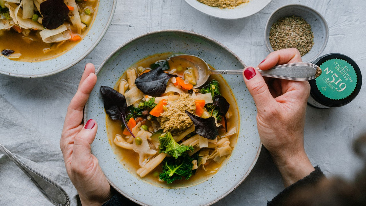 Gemüsesuppe mit Blattgemüse und Nudeln; eine Person hält einen Löffel in der Hand.