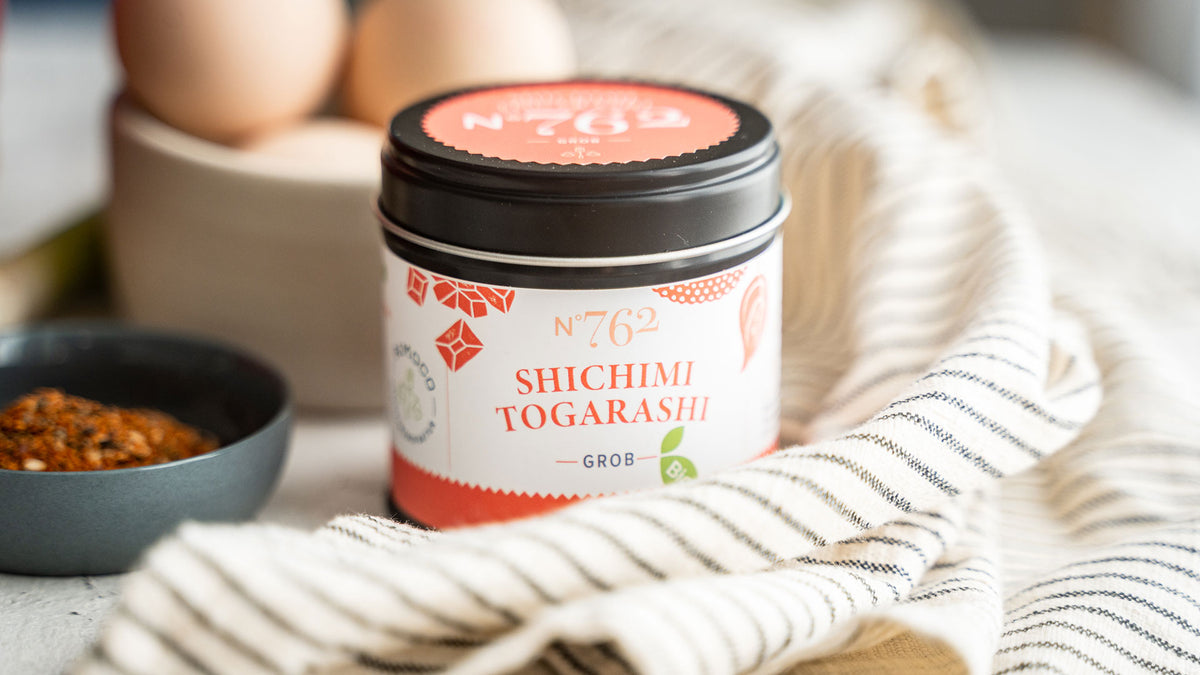 Ein Glas Shichimi Togarashi steht auf einem gestreiften Tuch neben einer Schale mit Chiliflocken.