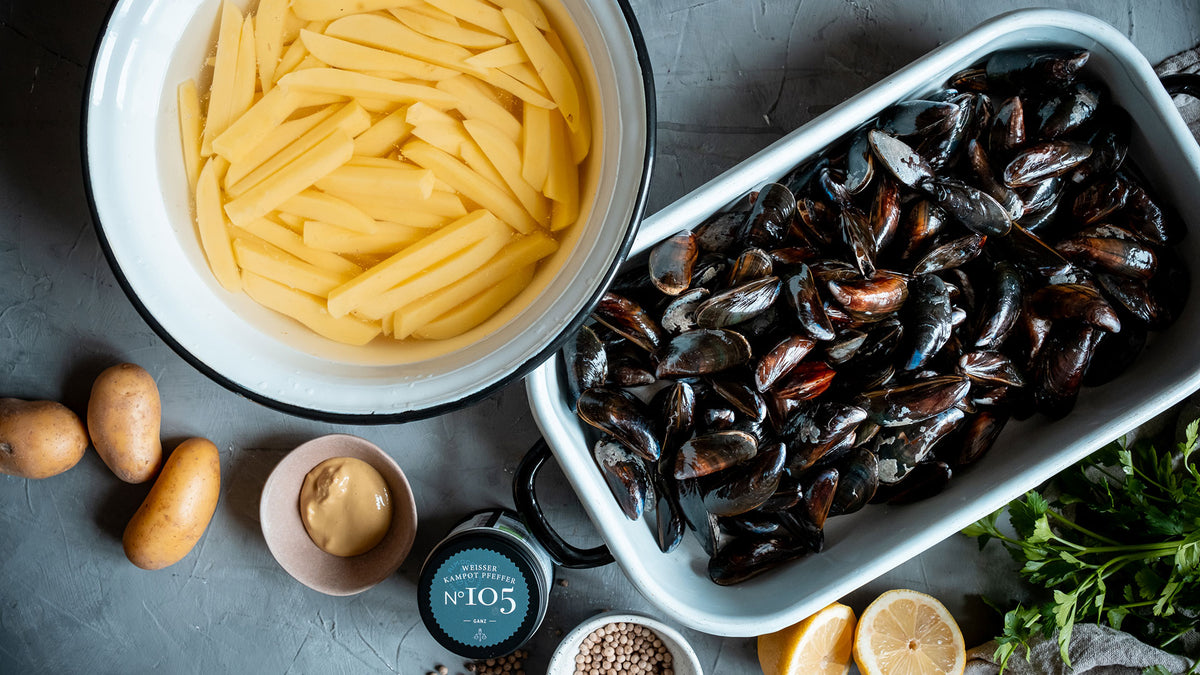 Geschnittene Kartoffeln in Wasser, Muscheln, Kräuter, Zitronenhälften und Gewürze auf einem Tisch.