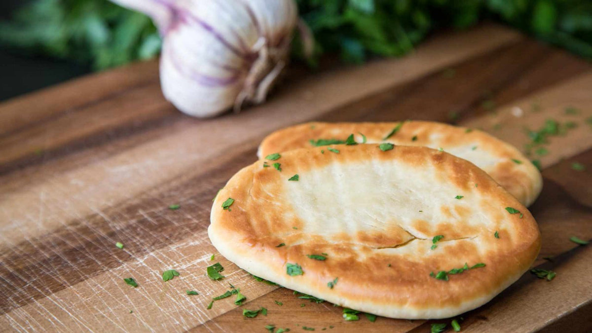Naanbrot mit gehackten Kräutern auf einem Holzbrett, im Hintergrund eine Knoblauchknolle.