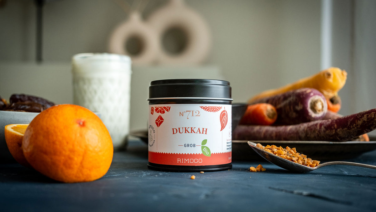 Auf einem Tisch steht ein Glas mit einer Dukkah-Gewürzmischung, daneben liegen Obst, Karotten und ein Löffel.