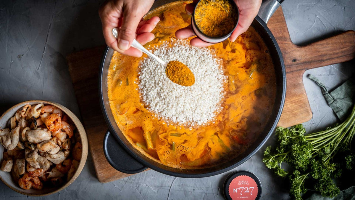 Reis in orangefarbener Brühe mit Garnelen, frischen Kräutern und Pilzen; eine Hand streut gelbes Gewürz darüber.