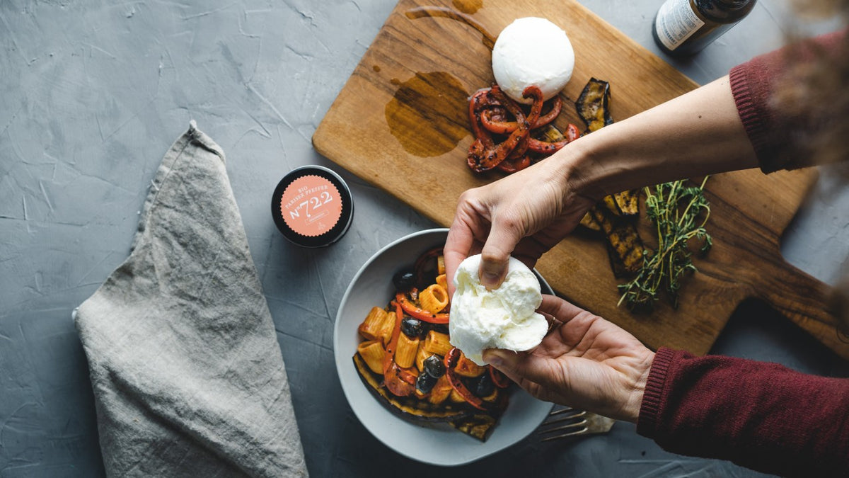 Zwei Hände reißen frischen Mozzarella über geröstetem Gemüse auf einem grauen Tisch mit einer Leinenserviette.