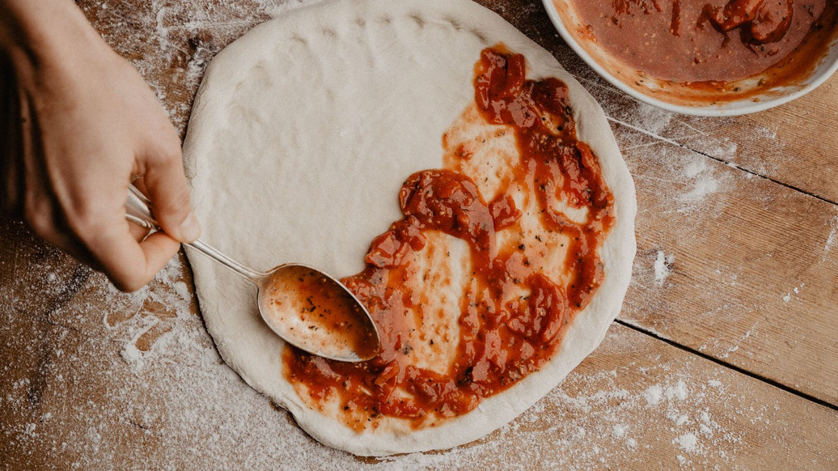 Eine Hand verteilt mit einem Löffel Tomatensauce auf dem Pizzateig, der auf einer Holzunterlage liegt.