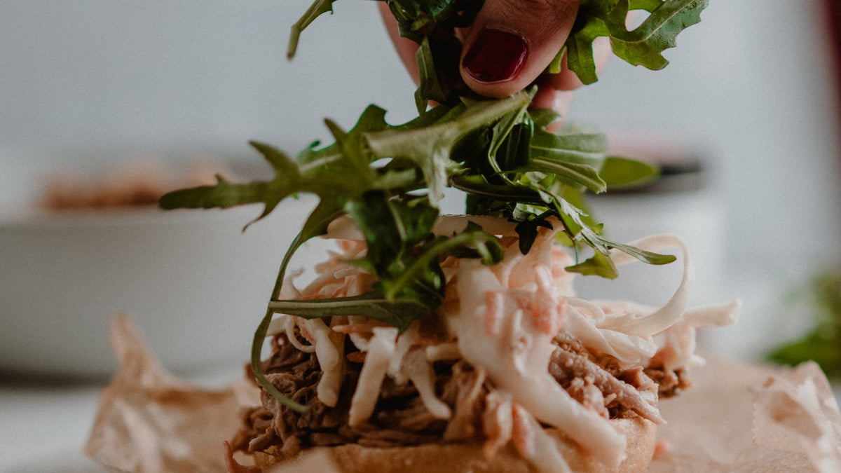 Jemand legt frischen Rucola auf ein Pulled-Pork-Sandwich mit Krautsalat.